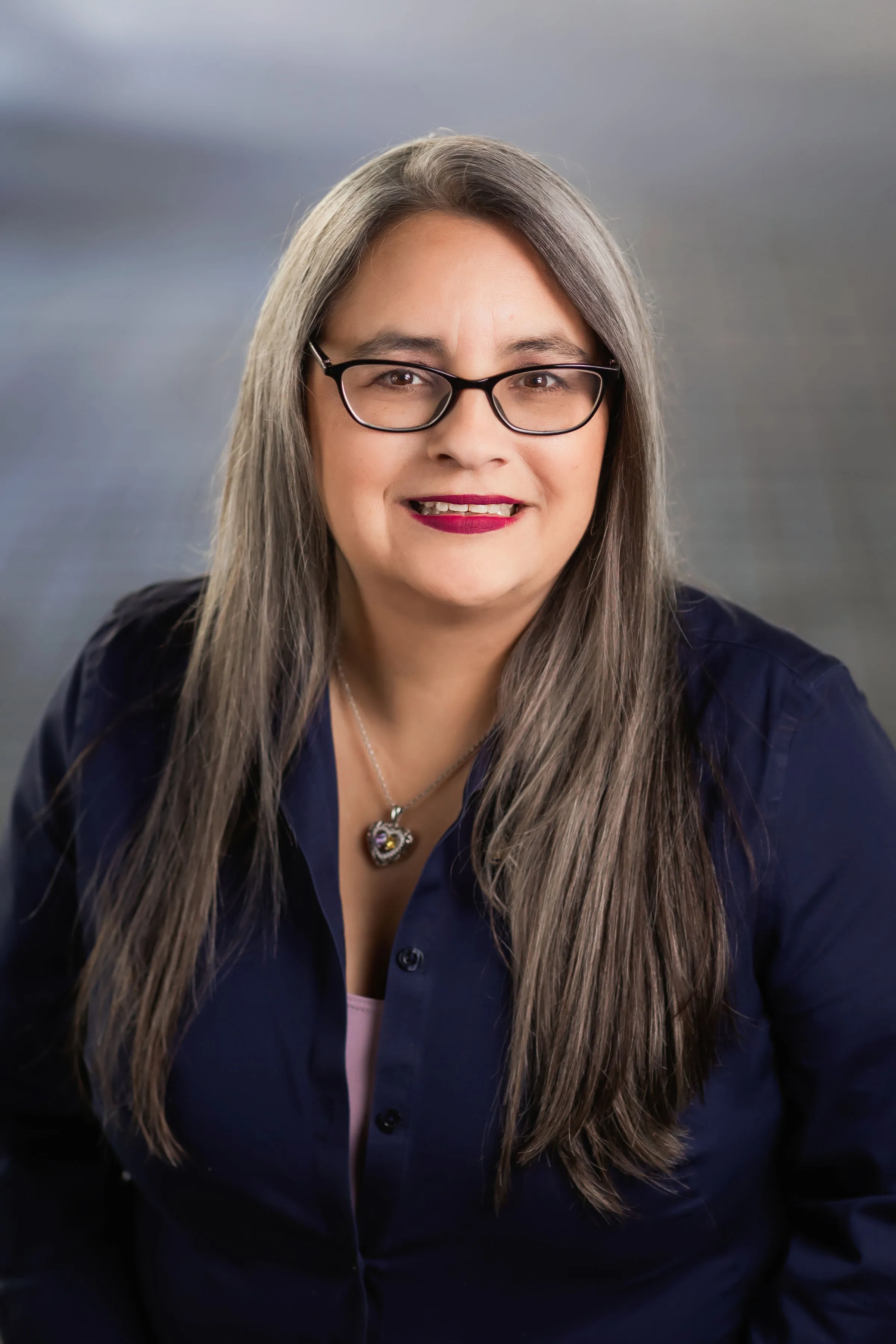 Professional woman with long gray hair and glasses, smiling confidently.